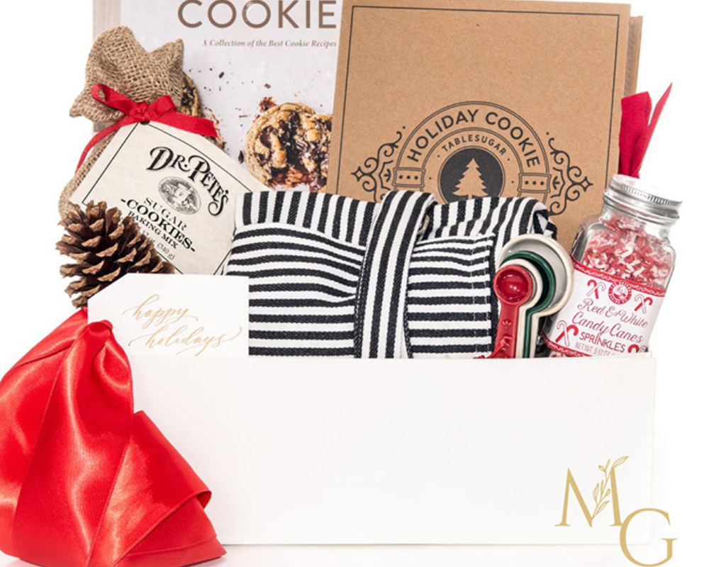 Holiday-themed cookie baking gift set featuring a cookbook titled “Let’s Eat Cookie,” recipe cards, striped kitchen towel, red measuring spoons, jar of candy cane sprinkles, cookie mix, pinecone, and a festive red ribbon, arranged in a white gift box with