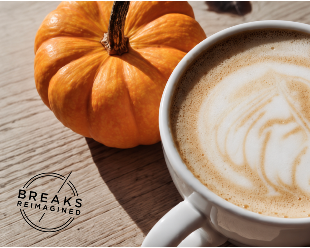 A steaming cup of coffee with latte art sits on a wooden table, with a mini pumpkin next to it, with a ‘Breaks Reimagined’ logo in the corner.
