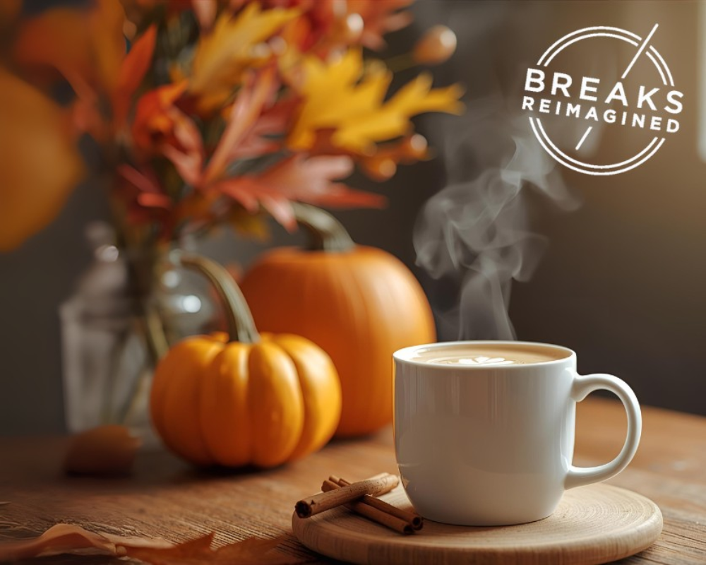 A steaming cup of coffee with latte art sits on a wooden coaster, surrounded by cinnamon sticks, pumpkins, and autumn leaves, with a 'Breaks Reimagined’ logo in the corner.