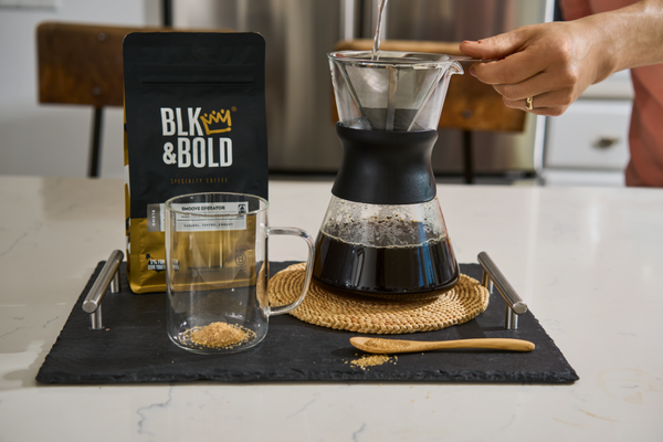Hand pouring hot water through a pour-over coffee maker next to a bag of BLK & Bold coffee and an empty glass mug on a counter.