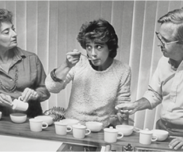 Black-and-white photo of three people seated at a table with several cups and bowls; the person in the center tastes something from a spoon while the two others watch attentively.