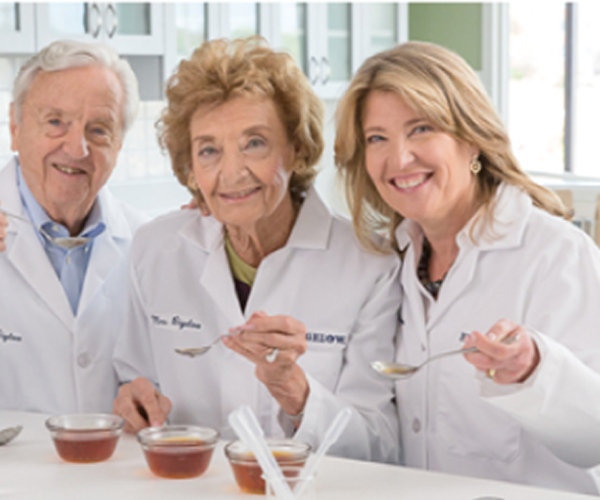 Three adults wearing white lab coats smile at the camera while holding spoons over small cups of what appears to be honey or another amber-colored food in a bright kitchen or lab setting.
